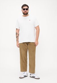 T-shirt blanc à manches courtes avec un petit logo rouge, associé à un pantalon en velours côtelé beige et des baskets blanches avec des rayures noires. Des lunettes de soleil noires sont portées.