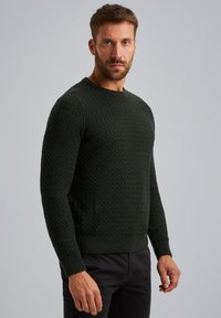 Donkergroen sweater met een gestructureerd diamantpatroon. Voorzien van een ronde halslijn en geribde manchetten en zoom. Draagt met donkere broek.
