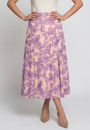 Donna che indossa una gonna midi rosa con stampa floreale viola, abbinata a décolleté beige con punta a punta, con le mani appoggiate sui fianchi, in piedi su sfondo grigio.