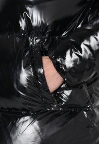 Černá péřová bunda s lesklým, reflexním povrchem, viditelným švem a rukou sahající do boční kapsy pro funkční detail.