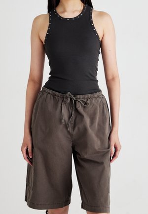 Femme portant un débardeur côtelé noir sans manches avec des détails cloutés et un short marron ample à cordon, sur un fond uni.