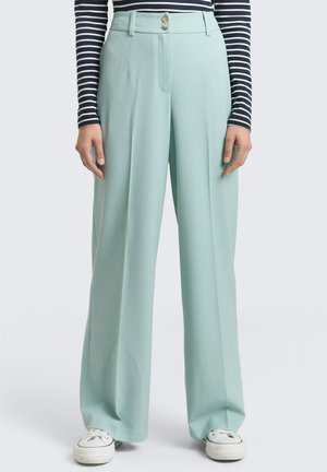 Frau trägt hellblaue, weit geschnittene Hosen mit doppeltem Knopfverschluss an der Taille, kombiniert mit einem langärmligen marineblau-weiß gestreiften Oberteil und weißen Sneakern.