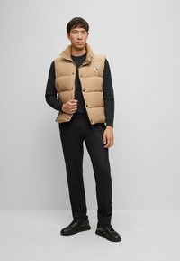 Beige gepolsterte Weste mit hohem Kragen, Knöpfen und Logo, getragen über einem schwarzen Langarmshirt und kombiniert mit schwarzen Hosen und Sneakers.