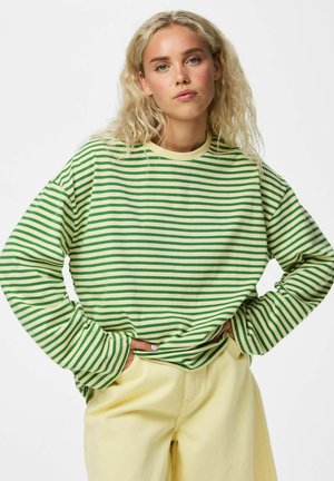 Jeune femme aux cheveux blonds bouclés portant un chemisier rayé vert et jaune à manches longues et un pantalon jaune clair, les mains dans les poches.