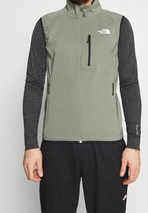 Man wearing light green sleeveless zip jacket with zippered chest and side pockets over a dark gray long-sleeve shirt and black pants.