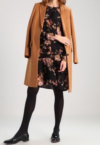 Brauner Wollmantel über einem schwarzen Blümchenkleid mit Rosa- und goldfarbenen Akzenten, kombiniert mit schwarzen Strumpfhosen und Lacklederschuhen.