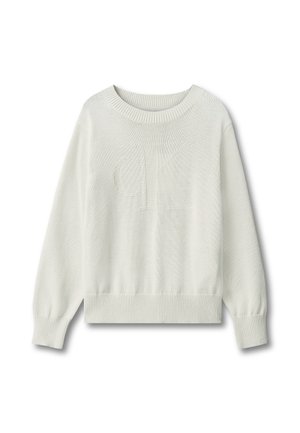 Dziecko z kręconymi blond włosami, ubrane w biały sweter, niebieskie dżinsy i czarne buty, stojące na jednolitym szarym tle.