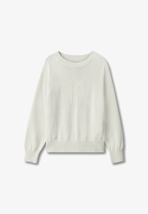 Barn med krøllet blondt hår iført en hvid sweater, blå jeans og sorte sko, stående foran en ensfarvet grå baggrund.