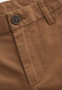 Pantalon en coton marron avec une texture lisse, doté d'une fermeture à bouton et de détails cousus le long de la taille et des poches.