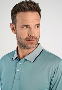 Uomo con capelli corti castani e barba che indossa una polo teal con un colletto grigio chiaro a trama, che guarda verso destra su uno sfondo bianco.