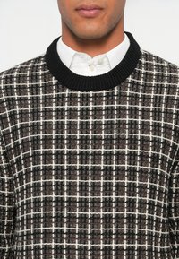 Άνδρας που φοράει ένα μαύρο και άσπρο καρό πλεκτό πουλόβερ πάνω από ένα λευκό πουκάμισο με κουμπιά, που φαίνεται από το λαιμό έως το μέσο του κορμού.