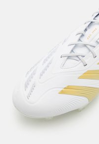Botas de fútbol blancas con parte superior texturizada, que presentan acentos dorados y cordones. Diseño ligero con estrías distintas para tracción.