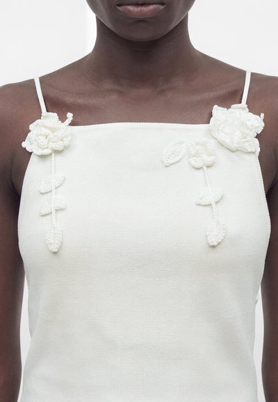 Άτομο με σκουρόχρωμη επιδερμίδα που φοράει λευκό πλεκτό αμάνικο τοπ με λεπτές τιράντες και τρισδιάστατες ανθισμένες εφαρμογές κοντά στους ώμους.