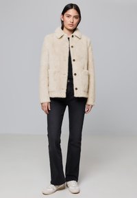 Veste crémeuse et moelleuse avec col, boutons devant et deux poches plaquées, assortie à un jean noir évasé et des baskets blanches.