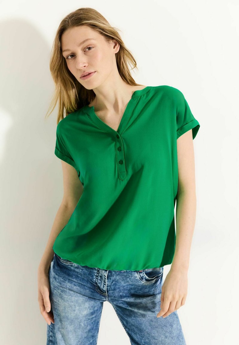Blusa verde a maniche corte con scollo a V e dettaglio dei bottoni. Realizzata in tessuto morbido, scivola dolcemente, abbinata a jeans blu sbiaditi.