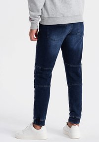 Jeans en denim bleu foncé avec une coupe fuselée, dotés de poches au dos et de poignets élastiques. Portés avec des baskets blanches et un sweatshirt gris.
