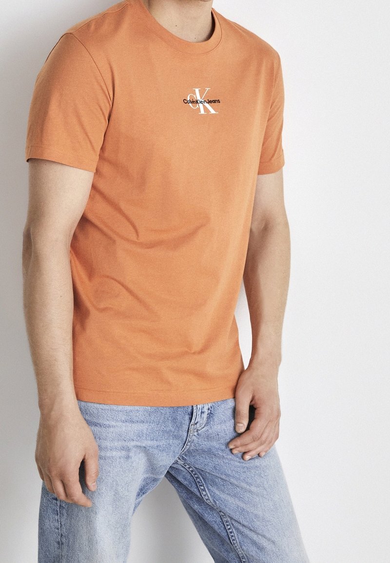 Oranžové bavlněné tričko s krátkými rukávy, kulatým výstřihem a černým logem Calvin Klein Jeans na hrudi, v kombinaci s světle modrými džínami.