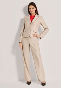 Beige op maat gemaakt pak met een getailleerde blazer, twee voorzakken en een bijpassende brede broek. Draagt een rode blouse eronder en zwarte hakken.