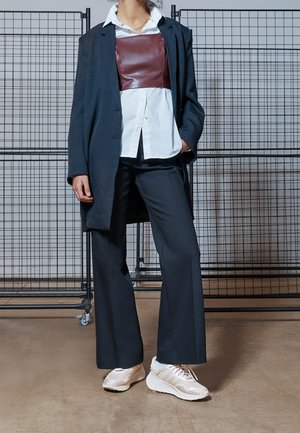 Tummanharmaa ylisuurikokoinen bleiseri, viininpunainen nahkainen korsetti valkoisen napillisen paidan päällä, leveälahkeiset tummat housut ja beiget urheilukengät.