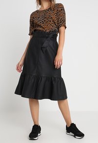 Top de bronce con estampado, falda corta de cuero ecológico negra con dobladillo con volantes, cintura ajustada con cinturón, combinada con zapatillas negras.