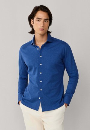 Jonge man met middellang donker haar, gekleed in een blauw overhemd met knopen en witte broek, staand met handen in de zakken.