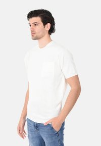 Camicetta bianca a maniche corte con scollatura rotonda e taschino sul petto, realizzata in un tessuto morbido e caratterizzata da una superficie liscia.