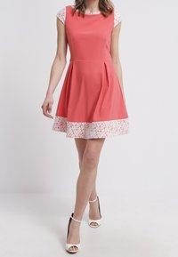 Kobieta w bezrękawnikowej koralowej sukience z białą koronką na rękawach i u dołu, ubrana w białe szpilki z kostką, stojąca na tle jednolitym.