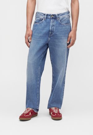 Personne portant un jean droit bleu clair, un haut blanc et des baskets rouges avec des lacets blancs, debout devant un fond uni.