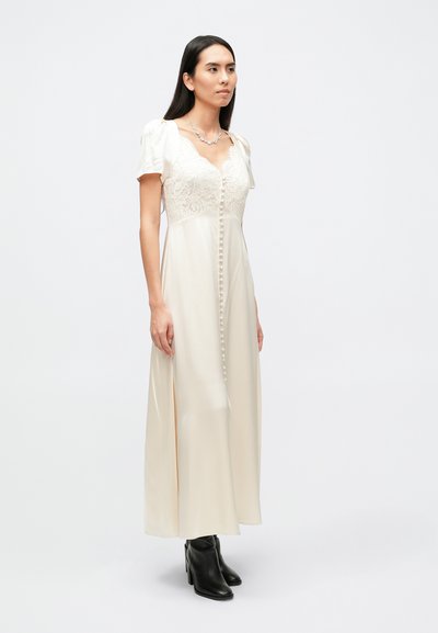 Ilga kreminė suknelė su nėrinių bodžiu, pūstomis rankovėmis ir sagų užsegimu priekyje, derinama su juodais kulkšnies aulinukais. Lygios tekstūros.