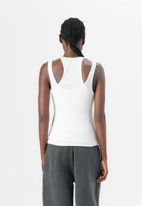White tank top with ribbed texture, featuring a scoop neckline and cut-out detail on the shoulders, paired with loose gray pants.