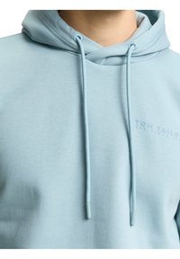 Sweat à capuche bleu clair avec cordons et logo "Tom Tailor" embossé subtil sur la poitrine, porté par une personne.