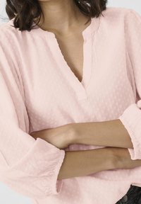 Lys rosa bluse med et struktureret mønster, V-hals og samlede, let puffede ærmer. Materialet ser blødt og letvægts ud.