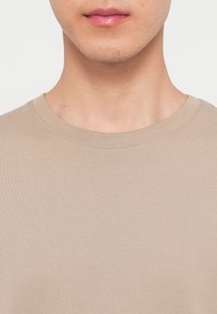 Close-up of a person’s lower face and neck wearing a beige crew-neck shirt against a plain background.