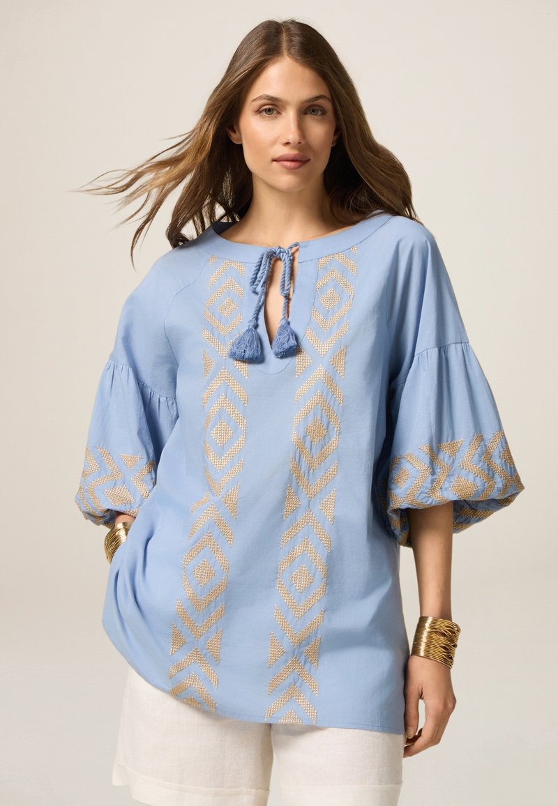 Blusa azzurro chiaro con maniche svasate, decorata con ricami geometrici dorati e chiusura con lacci e nappine al collo.