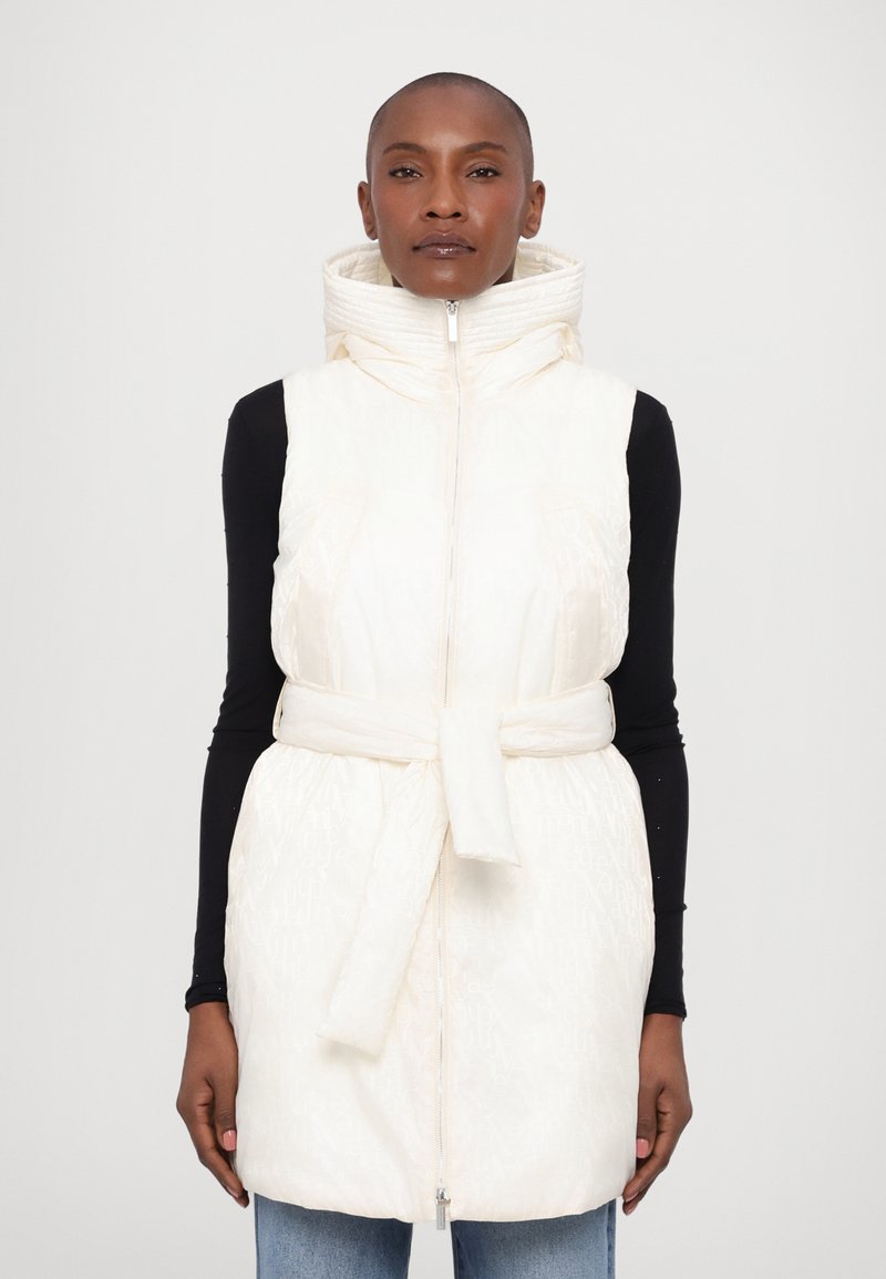 Padded vest in cream with a high collar, adjustable tie belt, two front pockets, and a zip closure. Textured fabric with subtle pattern.