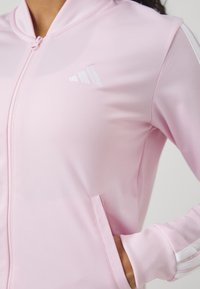 Rozā jaciņa ar rāvējslēdzēju, izgatavota no gludas materiāla, ar baltu Adidas logo un svītrām uz piedurknēm, ar sānu kabatām un ribotām piedurknēm.