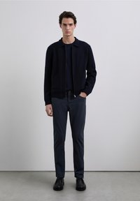 Black zip-up jacket with a collared neckline, paired with dark blue pants and black shoes. Smooth texture, minimalistic design, casual fit.