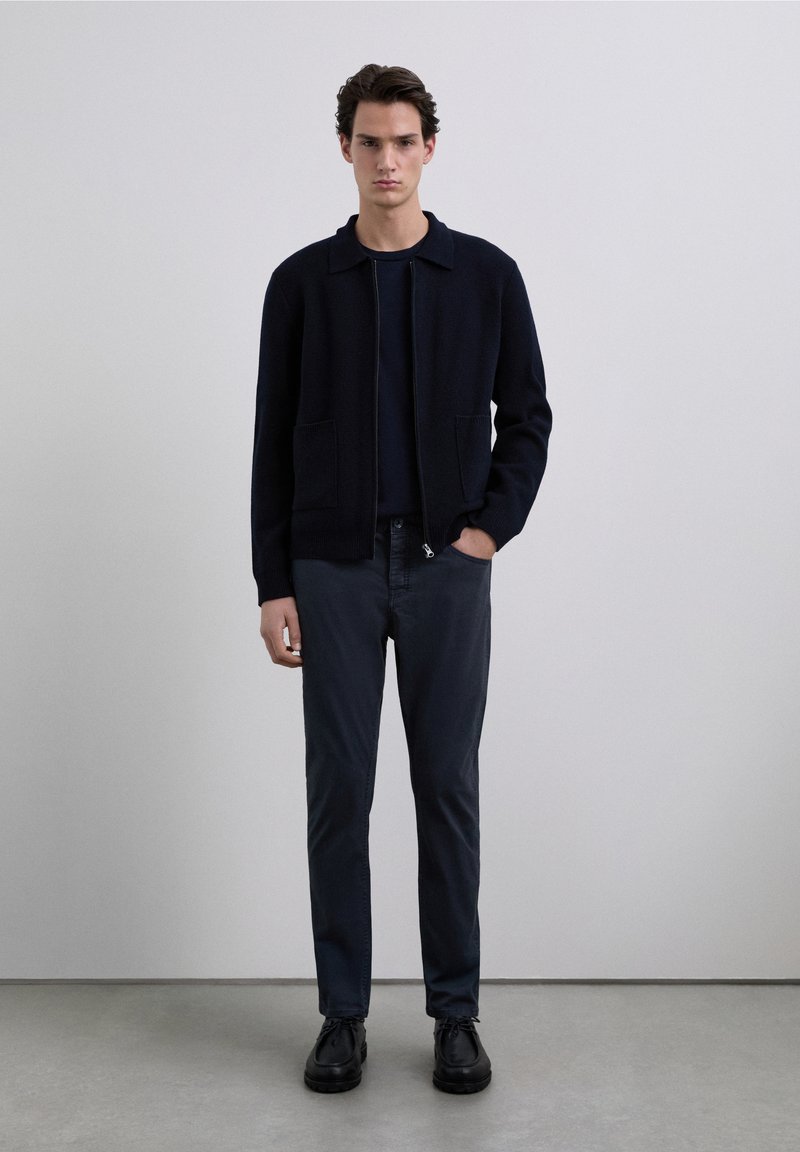 Black zip-up jacket with a collared neckline, paired with dark blue pants and black shoes. Smooth texture, minimalistic design, casual fit.