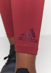 Czerwone legginsy sportowe o gładkiej fakturze, z ciemnoczerwonym logo Adidasa. Pojawia się widoczne przeszycie w mankiecie, a nogawki są dopasowane w kostce.