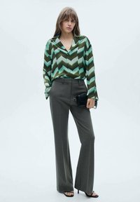 Blusa a righe chevron verde e nera con colletto, abbinata a pantaloni grigi a vita alta e borsa a tracolla nera. Tessuto morbido e vestibilità rilassata.