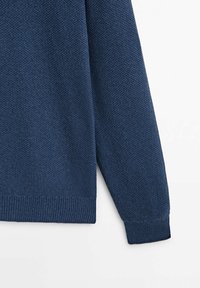 Pull en tricot bleu marine avec un motif texturé, muni de poignets et d'un ourlet côtelés, présenté partiellement, mettant l'accent sur la manche et le corps.