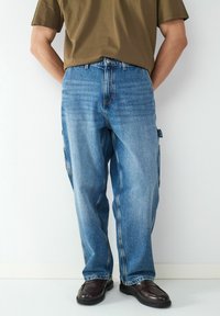 Jean en denim bleu avec une coupe décontractée, délavé clair et coutures visibles. Assorti avec une chemise à manches courtes marron et des chaussures foncées.