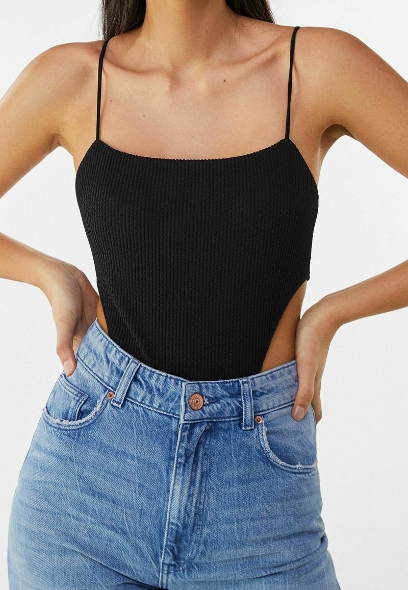 Femme portant un haut noir texturé sans manches à fines bretelles et un jean taille haute bleu clair, les mains sur les hanches.