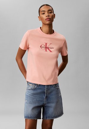 Mujer con camiseta rosa claro con el logo de Calvin Klein y pantalones cortos vaqueros, de pie con las manos detrás de la espalda contra un fondo gris liso.