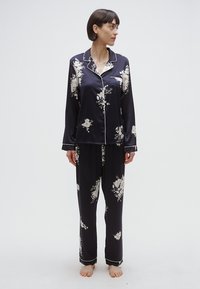 Ensemble de pyjama en satin bleu marine, présentant des motifs floraux en blanc et jaune, avec un liseré blanc contrastant sur le col et les poignets. Manches longues, pantalon ample.