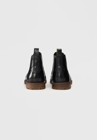 Botas de tobillo de cuero negro con tiradores amarillos, suelas de goma y acentos de logo hexagonal. Forma compacta con textura suave y suelas marrones.
