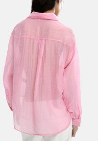 Lekka różowa koszula z przezroczystej tkaniny, z długimi rękawami i kołnierzem. Posiada mankiety na guziki oraz luźny krój z detalem w postaci plisek z tyłu.