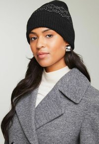 Gorro de lana negro con patrón geométrico y acentos plateados, combinado con un abrigo de lana gris y un suéter de cuello vuelto crema. Texturas suaves y líneas limpias.