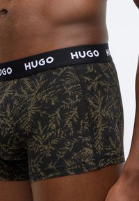Čierne boxerky s olivovo zeleným vzorom listov, s čiernym pásom, na ktorom je biely nápis "HUGO". Hladká textúra a priliehavý strih.