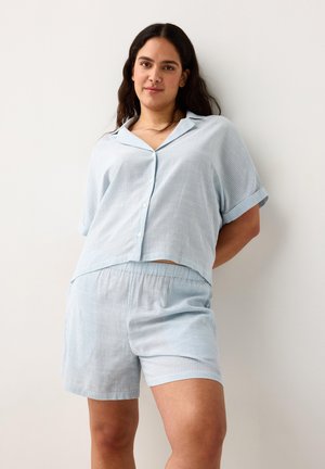 Kvinde med langt mørkt hår iført lyseblå stribet kortærmet skjorte med knapper og matchende shorts, stående mod en hvid væg.